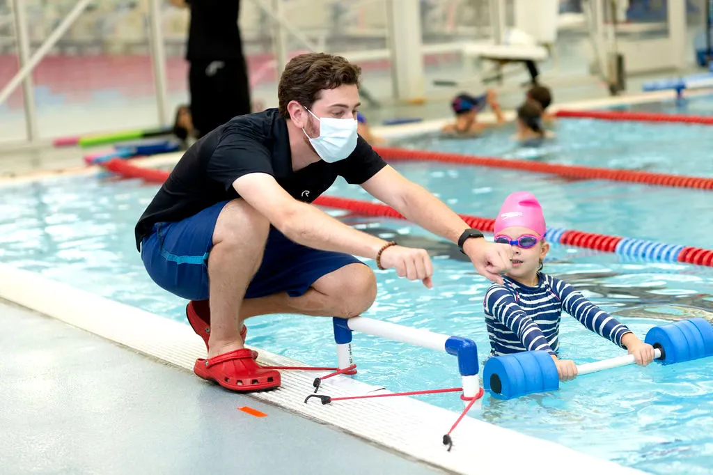 Coach instructing a young swimmer in a pool while wearing a mask.