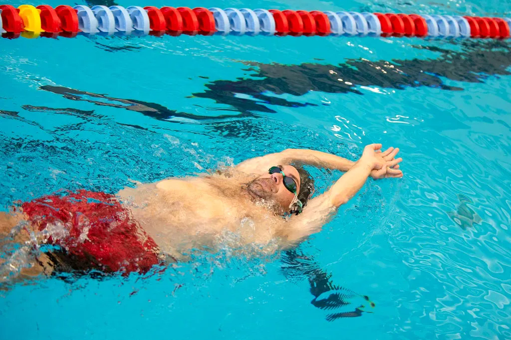 Swimmer in goggles and red swim trunks practicing backstroke in pool.